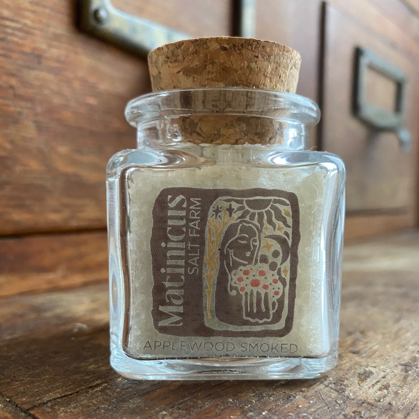 1.4 ounce glass jar containing applewood smoked sea salt with cork lid on a wooden surface