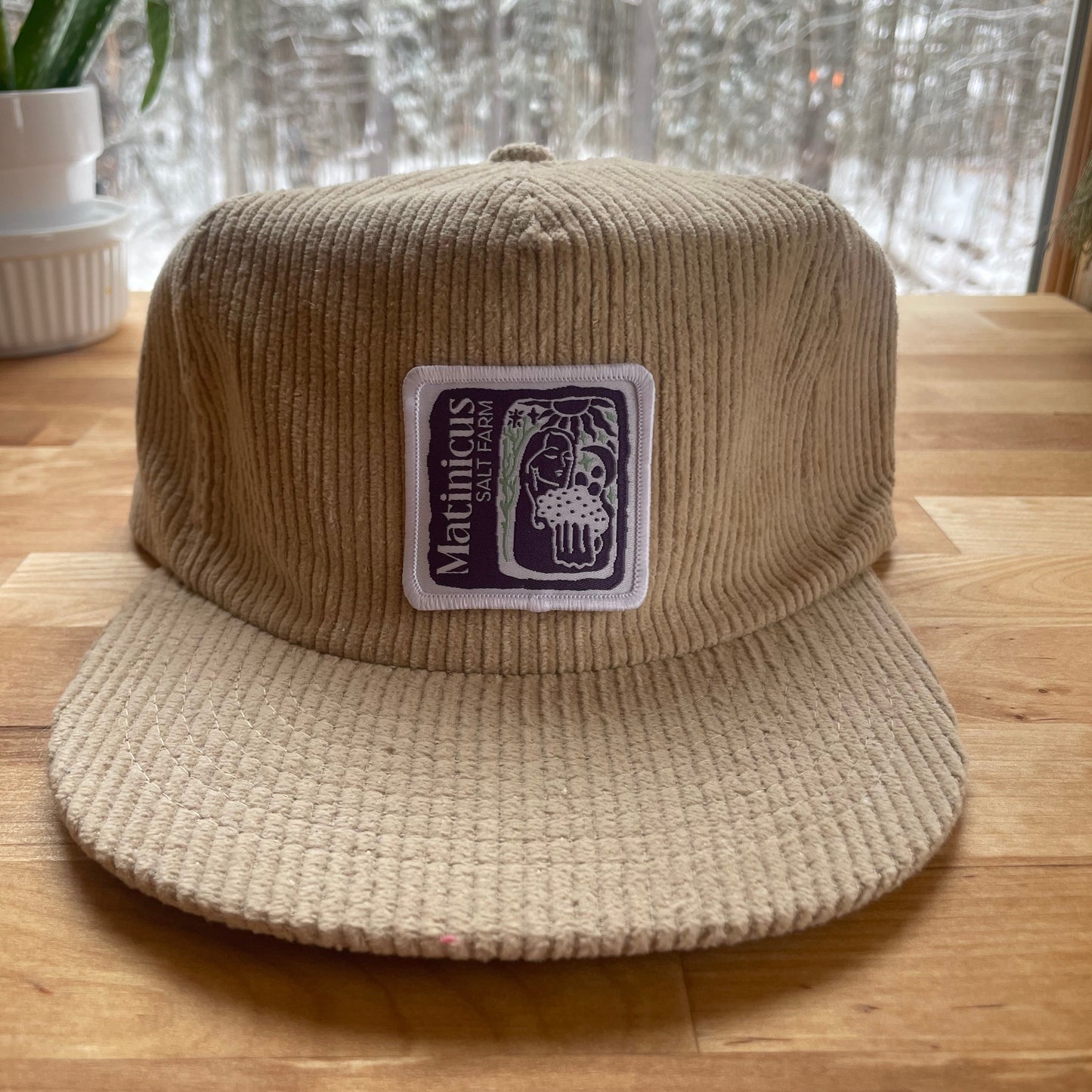 Beige corduroy cap with an indigo logo patch on a wooden surface