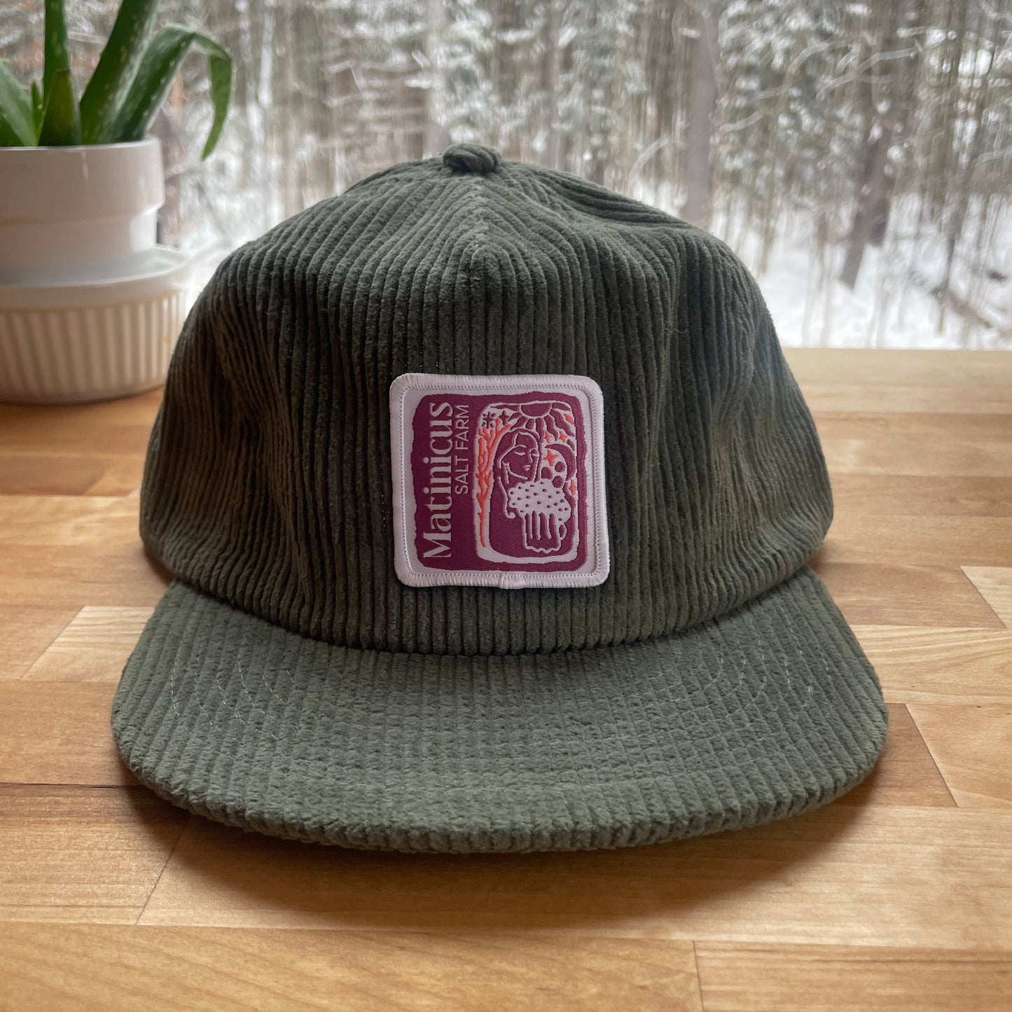 Green corduroy cap with a plum logo patch on a wooden surface