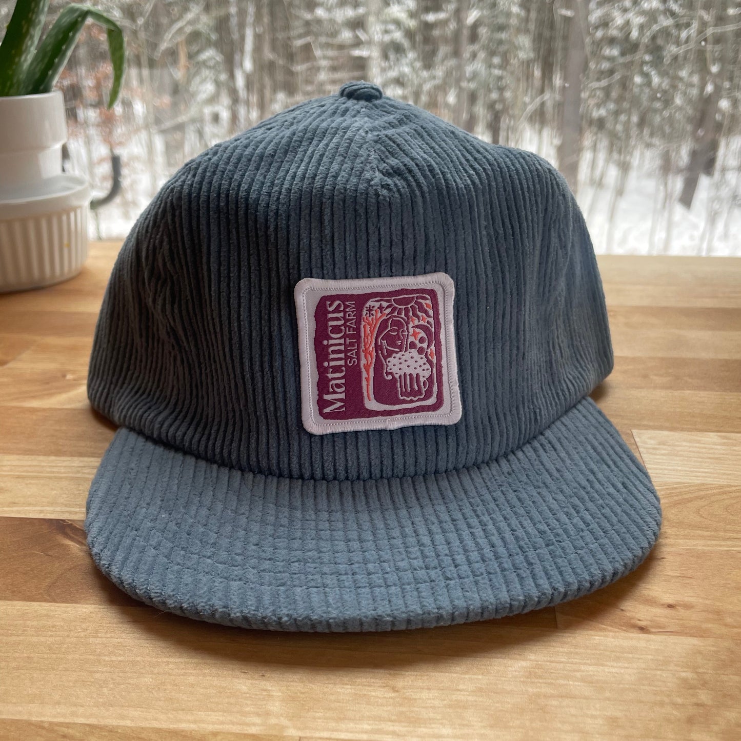 Blue corduroy cap with a plum logo patch on a wooden surface