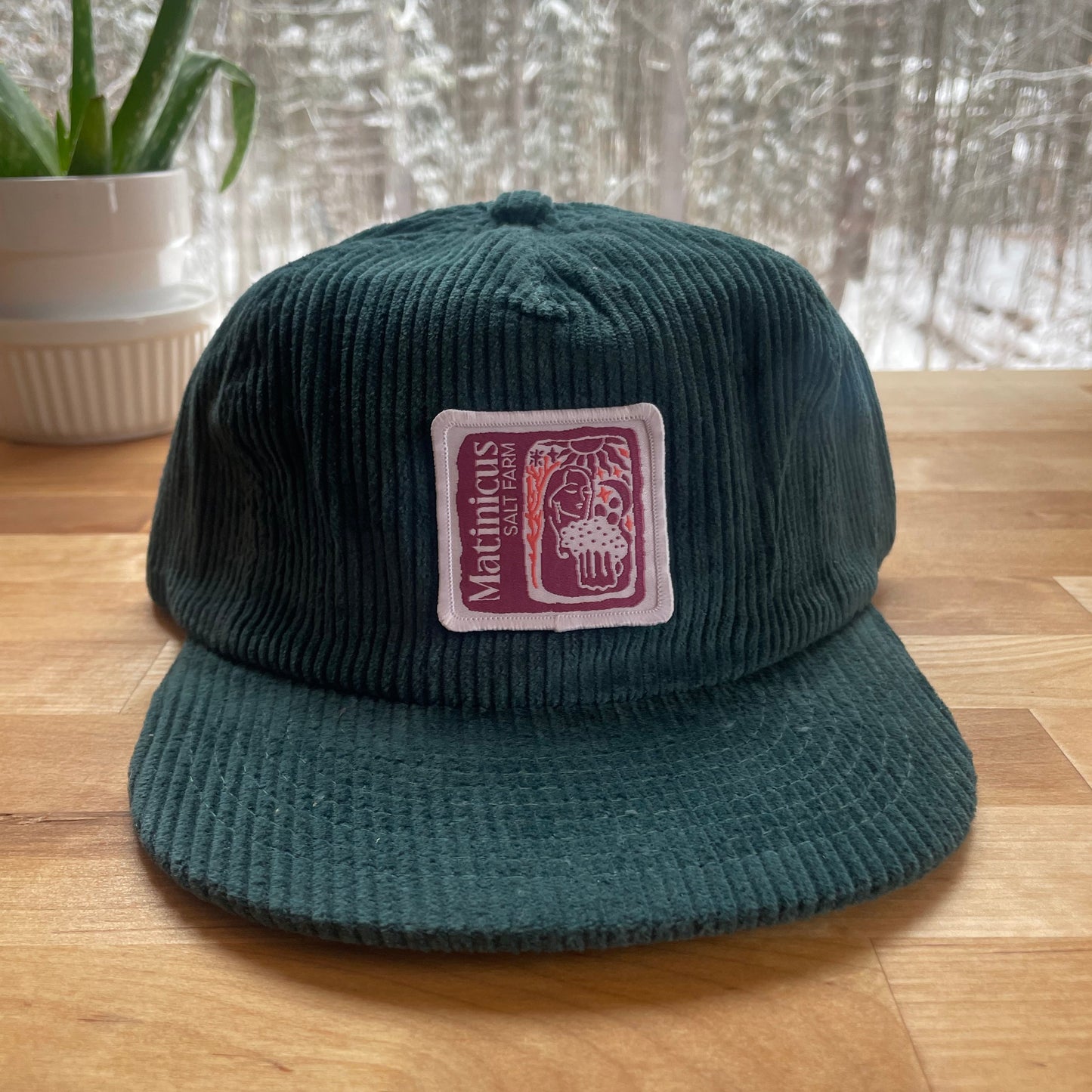 Dark teal corduroy cap with a plum logo patch on a wooden surface