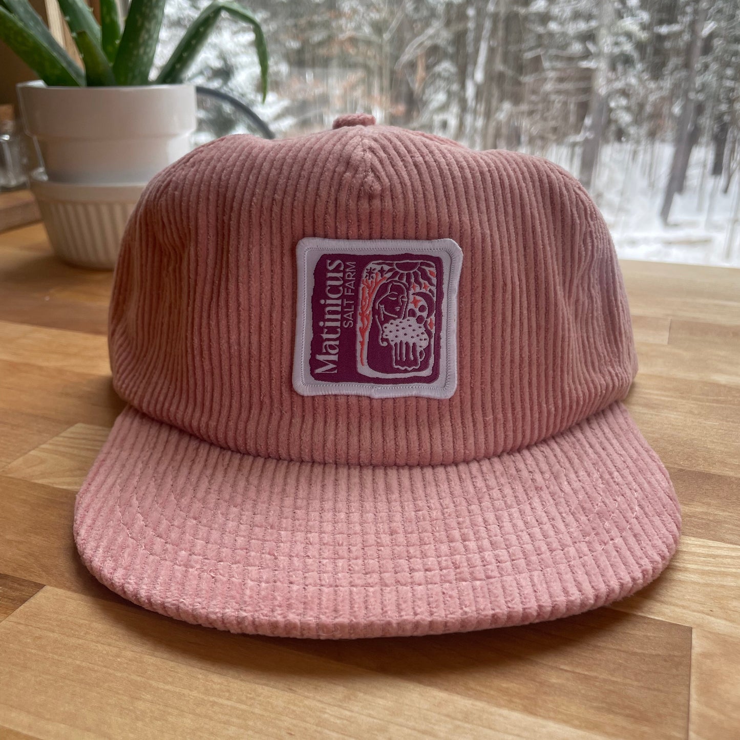 Pink corduroy cap with a plum logo patch on a wooden surface