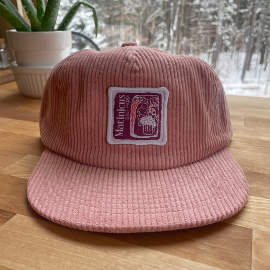 Pink corduroy cap with a plum logo patch on a wooden surface