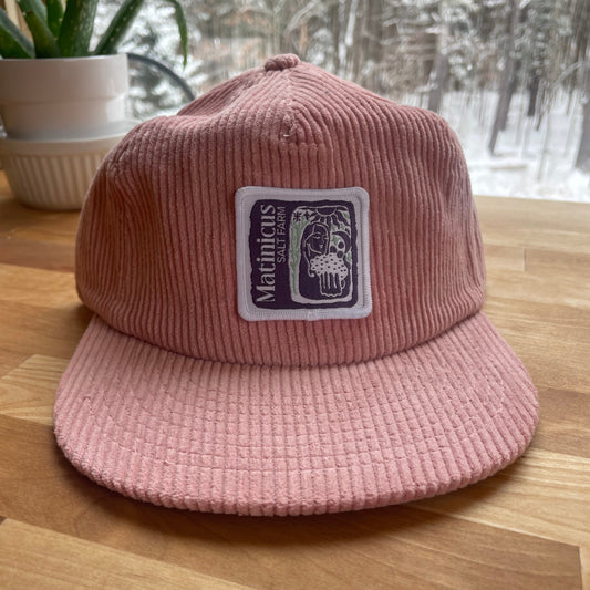 Pink corduroy cap with an indigo logo patch on a wooden surface