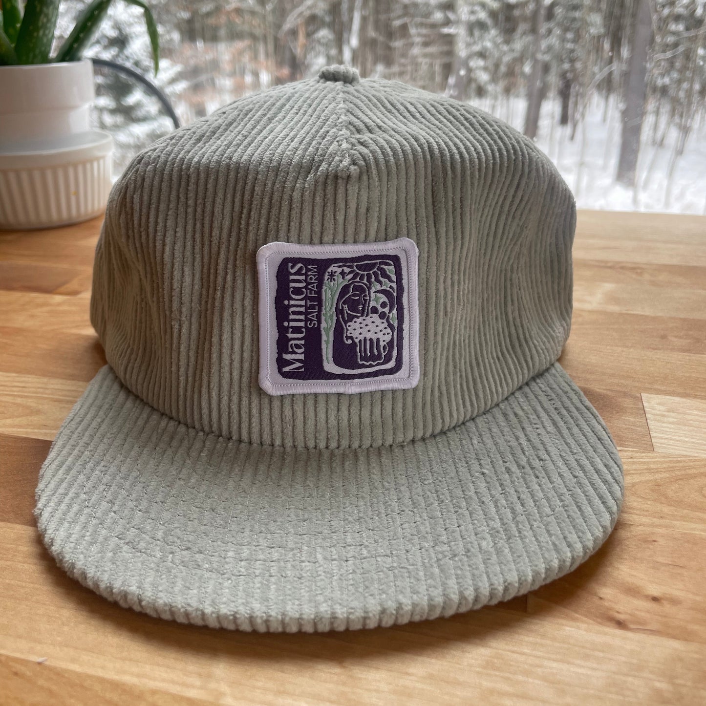 Gray corduroy cap with an indigo logo patch on a wooden surface