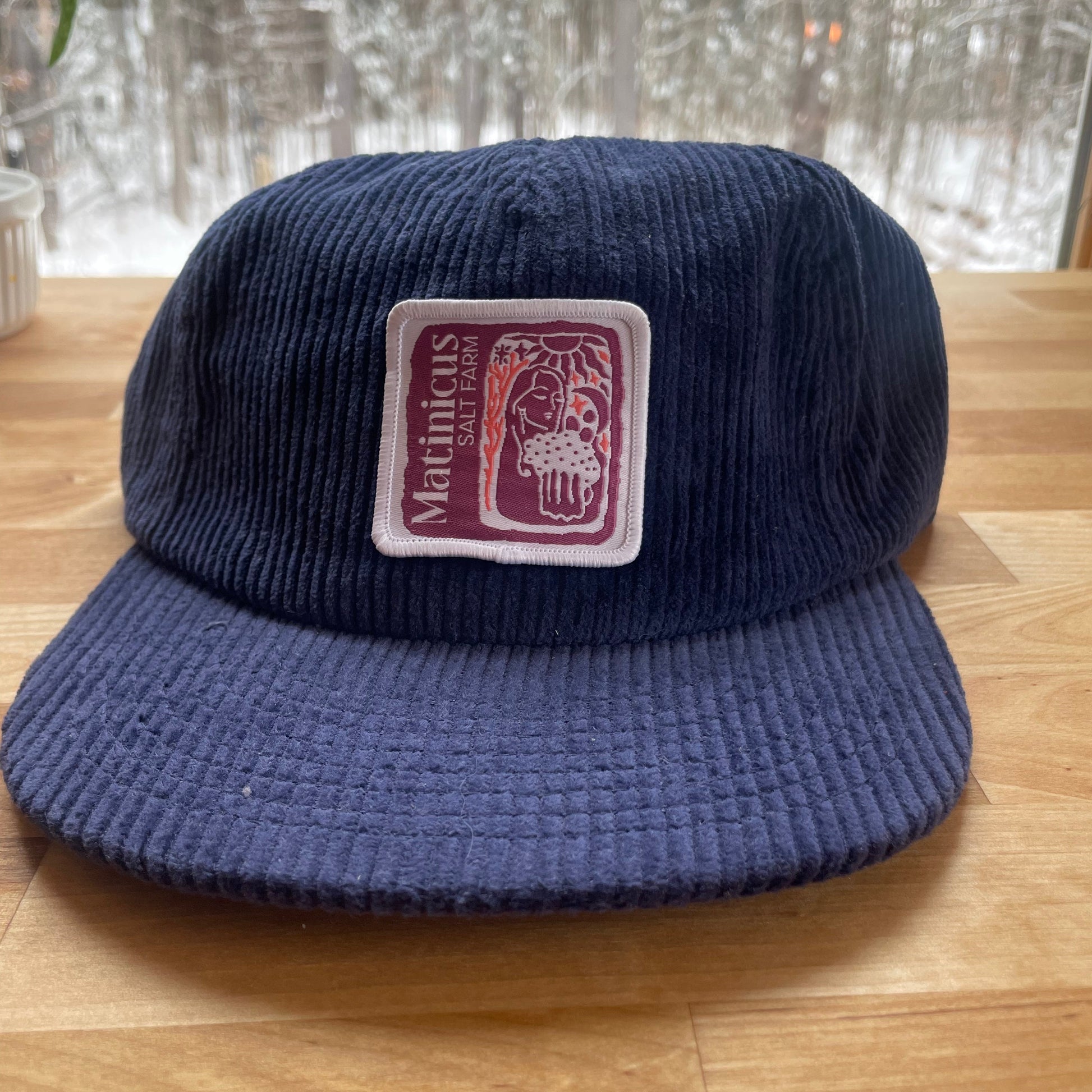 Navy blue corduroy cap with a plum logo patch on a wooden surface
