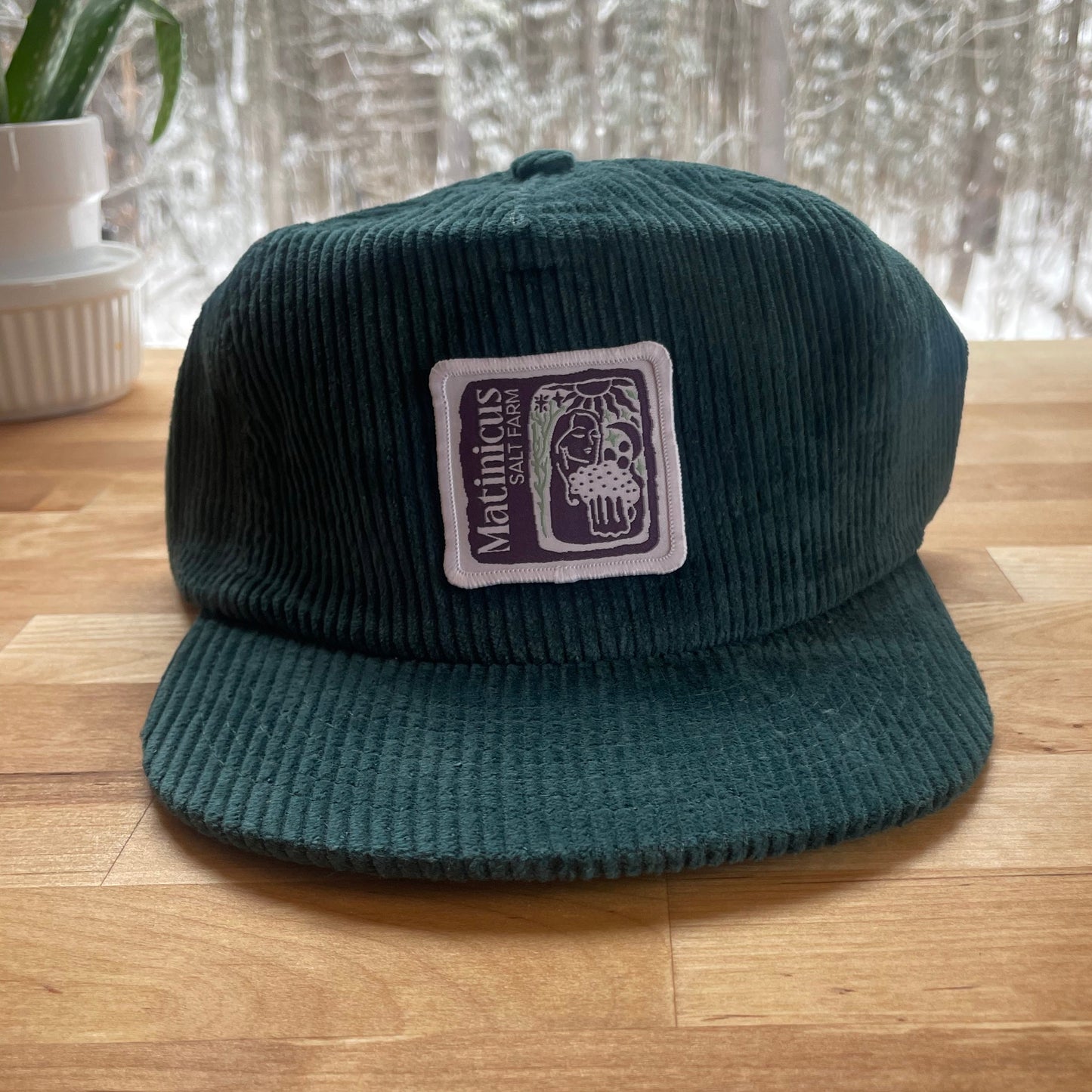 Dark teal corduroy cap with an indigo logo patch on a wooden surface