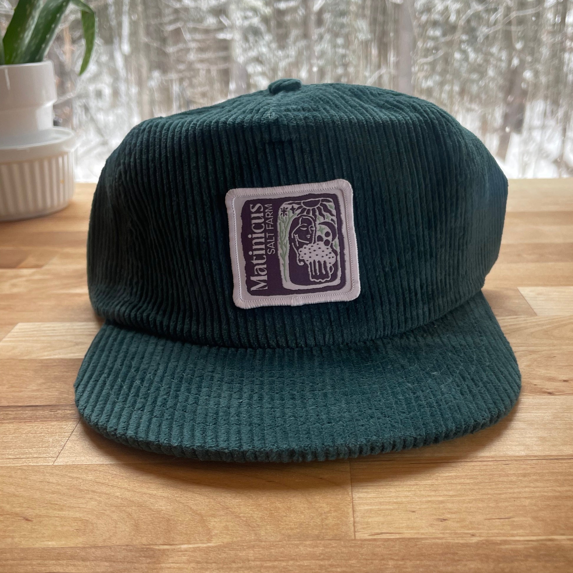 Dark teal corduroy cap with an indigo logo patch on a wooden surface